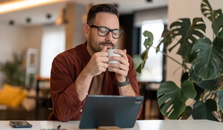 man drinking coffee in front of laptop with eyes closed
