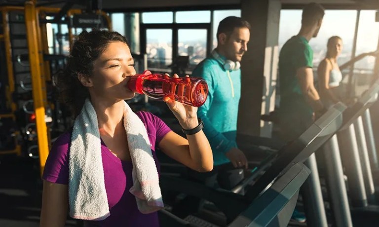woman drinking red water bottle while running on treadmill